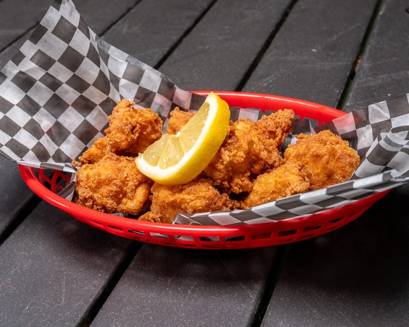 Golden fried chicken bites with a lemon wedge in a red basket lined with checkered paper.