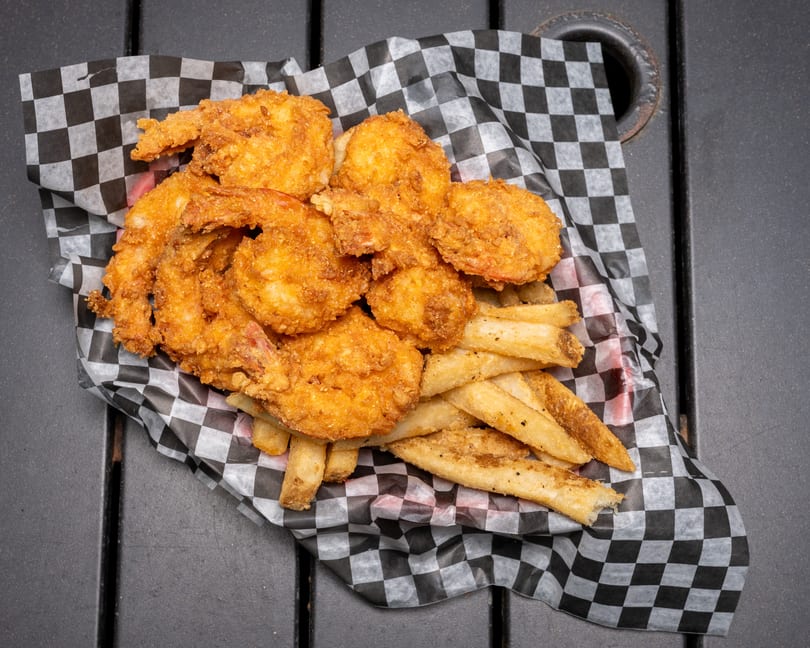 Golden-brown fried shrimp and seasoned french fries in a checkered paper-lined basket on a dark table.