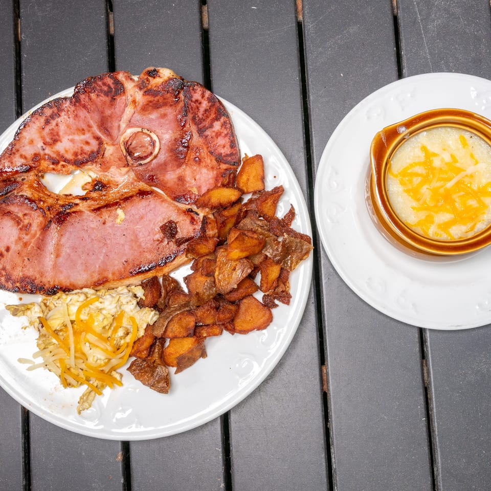 A breakfast plate with grilled ham, scrambled eggs, home fries, and a side of cheesy grits.