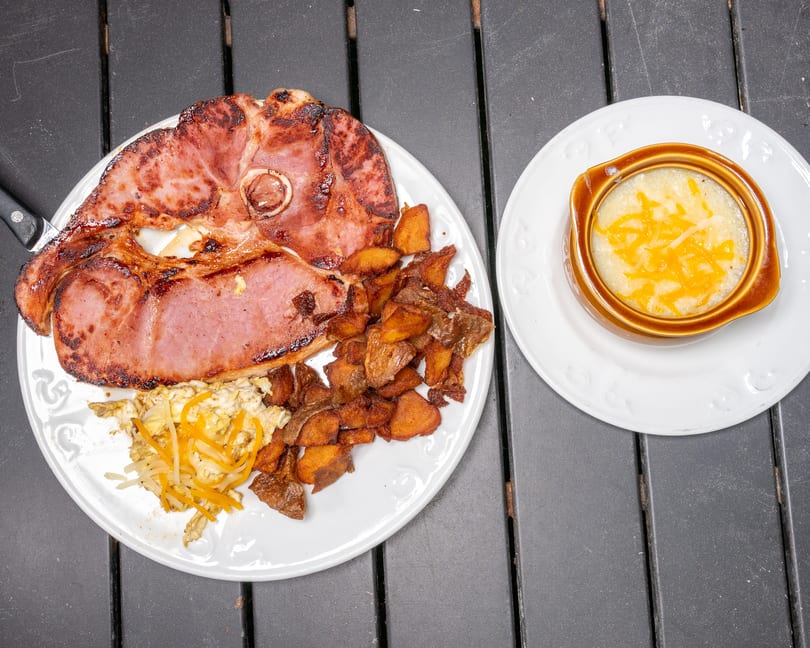 A breakfast plate with grilled ham, scrambled eggs, home fries, and a side of cheesy grits.