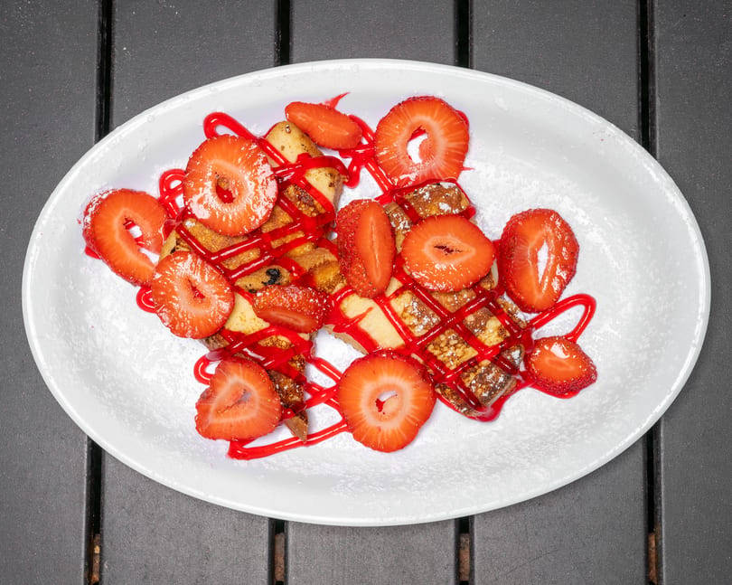 French toast sticks with powdered sugar, strawberry slices, and red syrup on a white plate.