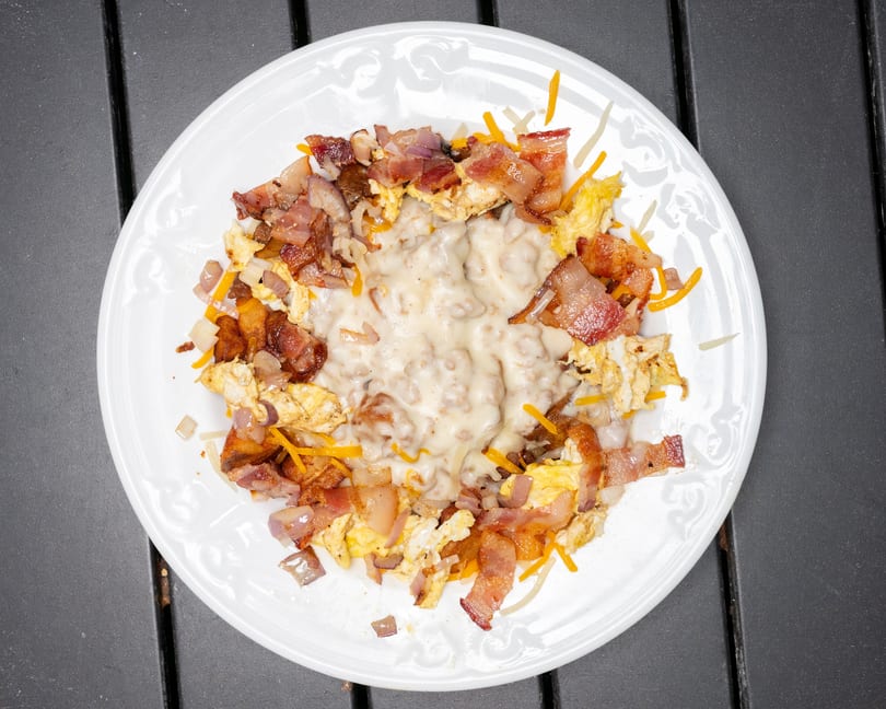 A breakfast skillet with scrambled eggs, bacon, onions, cheese, and gravy on a white plate.