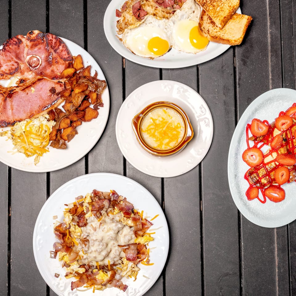 An overhead shot of five breakfast plates, including ham, eggs, bacon, grits, and strawberry French toast.