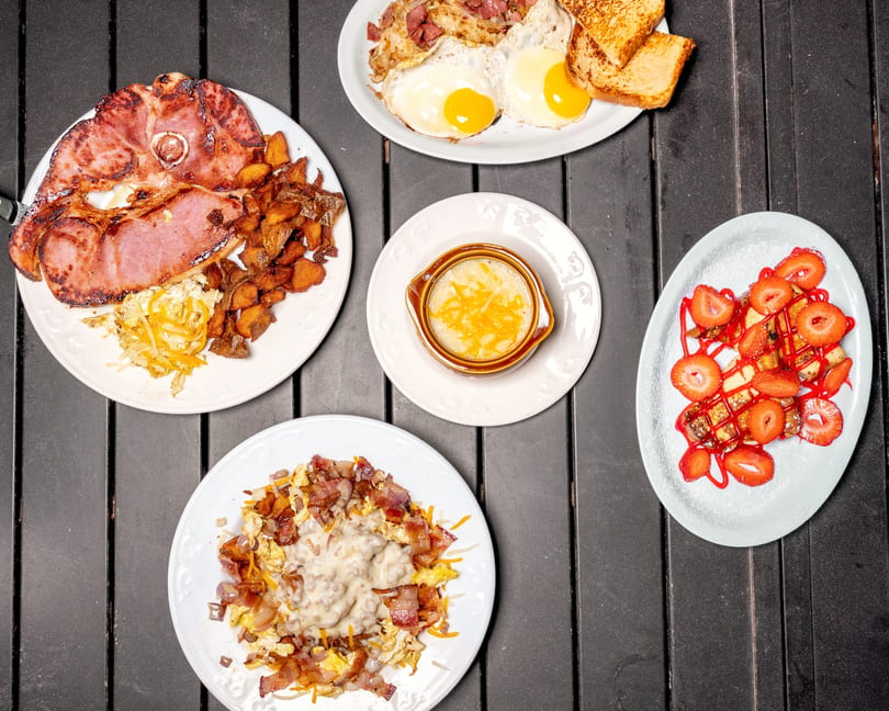 An overhead shot of five breakfast plates, including ham, eggs, bacon, grits, and strawberry French toast.