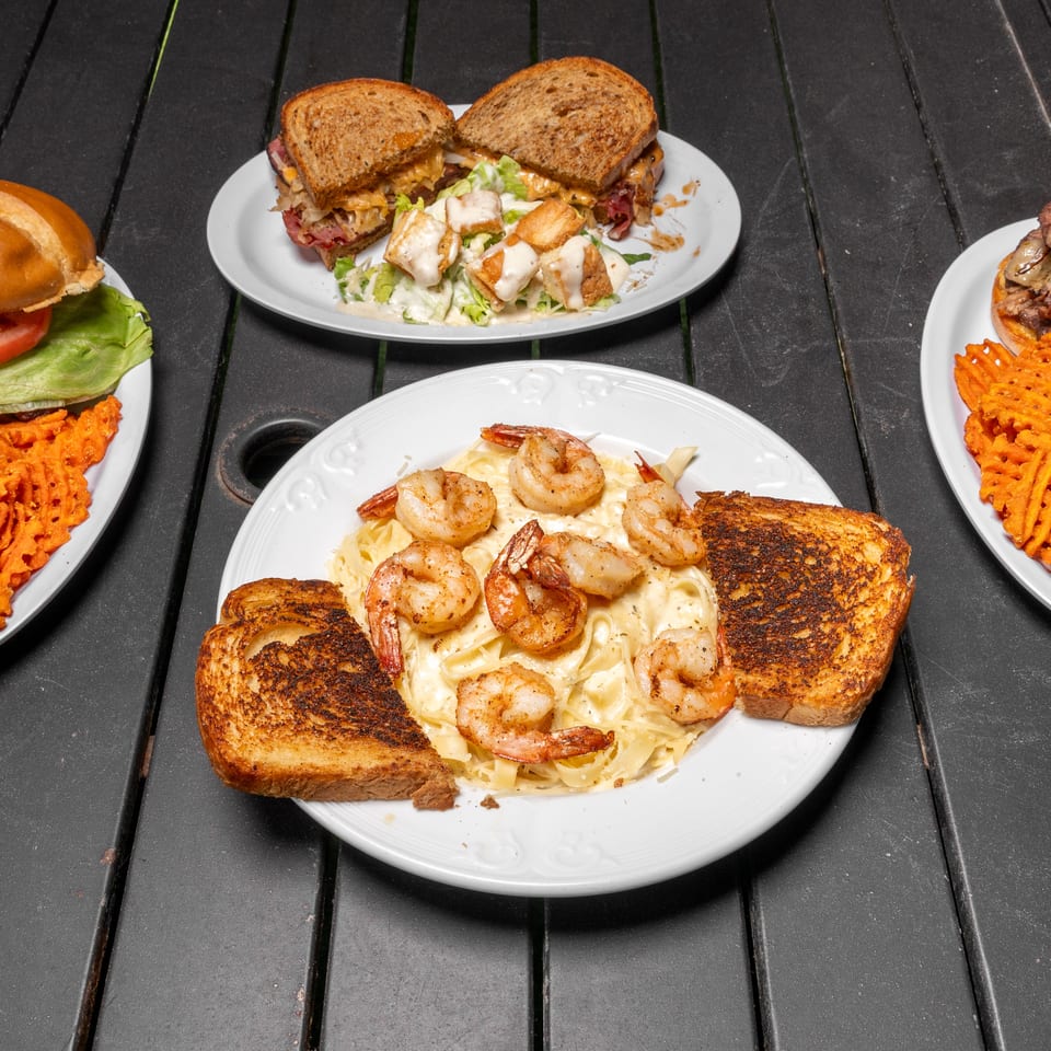 Four plates of food on a dark wooden table, including sandwiches, shrimp pasta, and burgers.