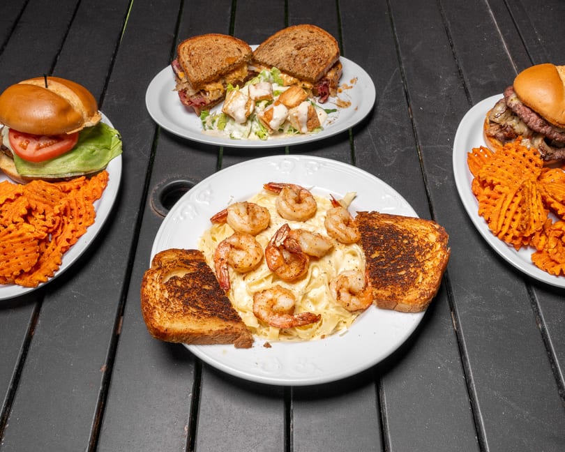 Four plates of food on a dark wooden table, including sandwiches, shrimp pasta, and burgers.
