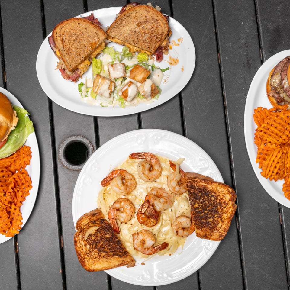 Overhead view of four plates of food, including sandwiches, burgers with waffle fries, and shrimp pasta.