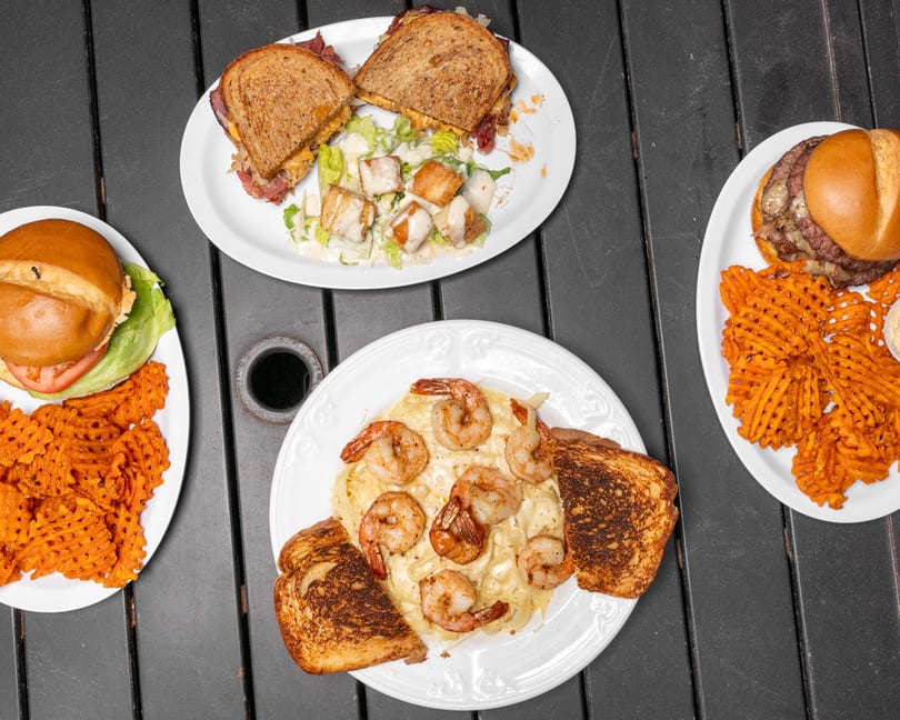 Overhead view of four plates of food, including sandwiches, burgers with waffle fries, and shrimp pasta.