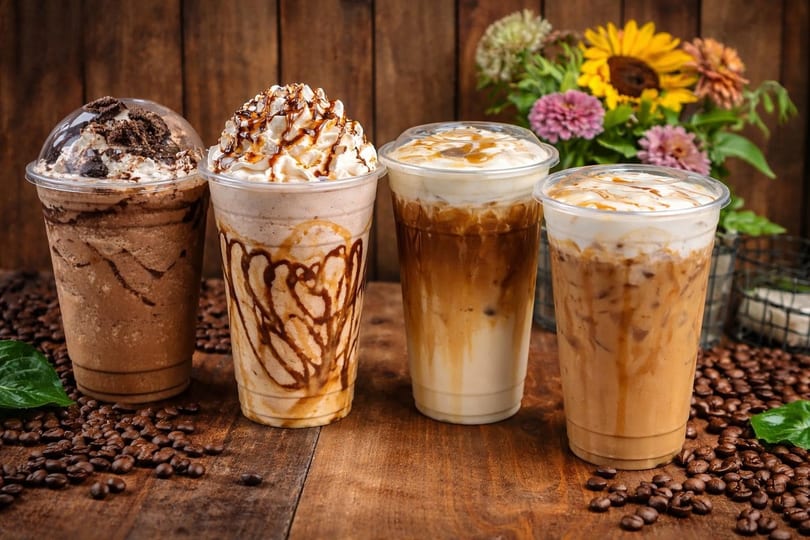 Four iced coffee drinks on a wooden table with coffee beans and flowers in the background.